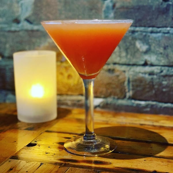 A vibrant orange cocktail in a martini glass next to a glowing candle on a wooden table.