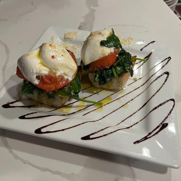 Two bruschetta pieces topped with burrata, tomatoes, and spinach, drizzled with balsamic glaze.