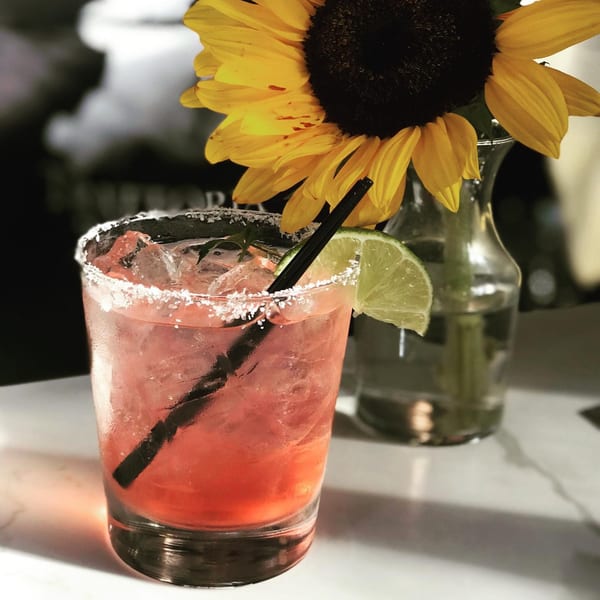 A vibrant red cocktail with a salted rim, lime wedge, and black straw, next to a sunflower.