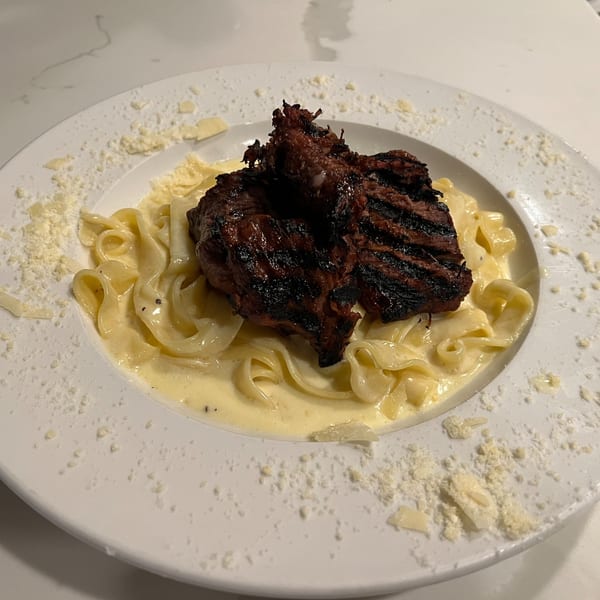 Grilled steak atop creamy fettuccine pasta, garnished with grated Parmesan cheese on a white plate.