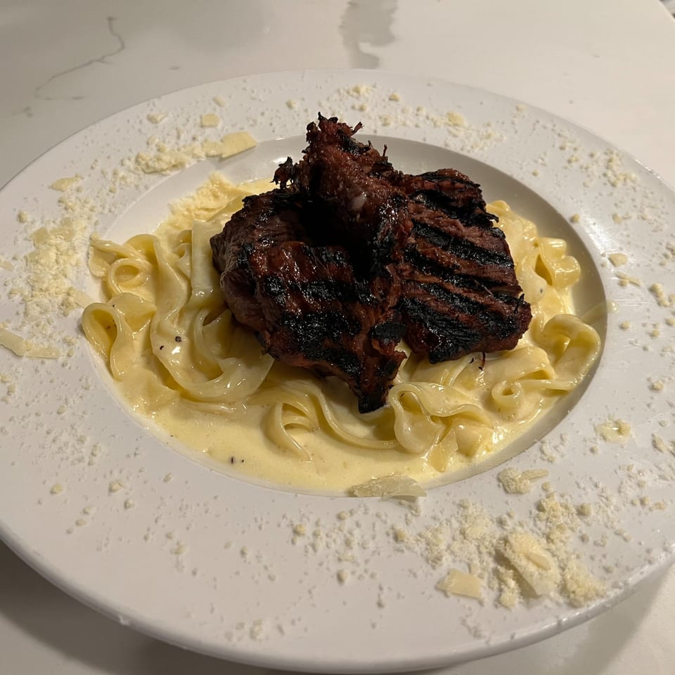 Grilled steak atop creamy fettuccine pasta, garnished with grated Parmesan cheese on a white plate.