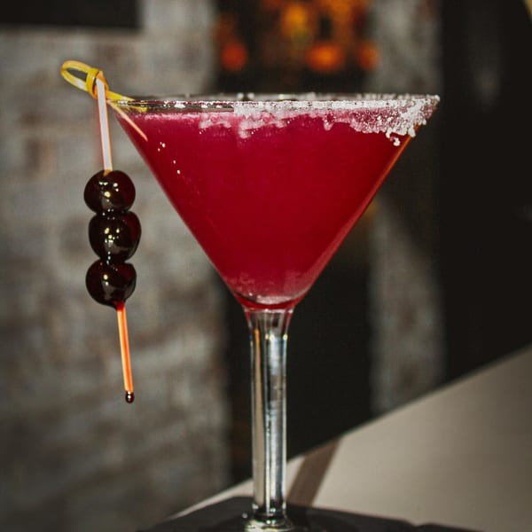 A vibrant red martini with a sugar-rimmed glass and three dark cherries on a pick.