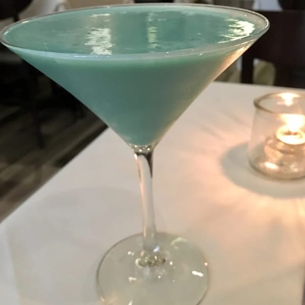 A vibrant blue-green martini in a stemmed glass sits on a white tablecloth next to a flickering candle.