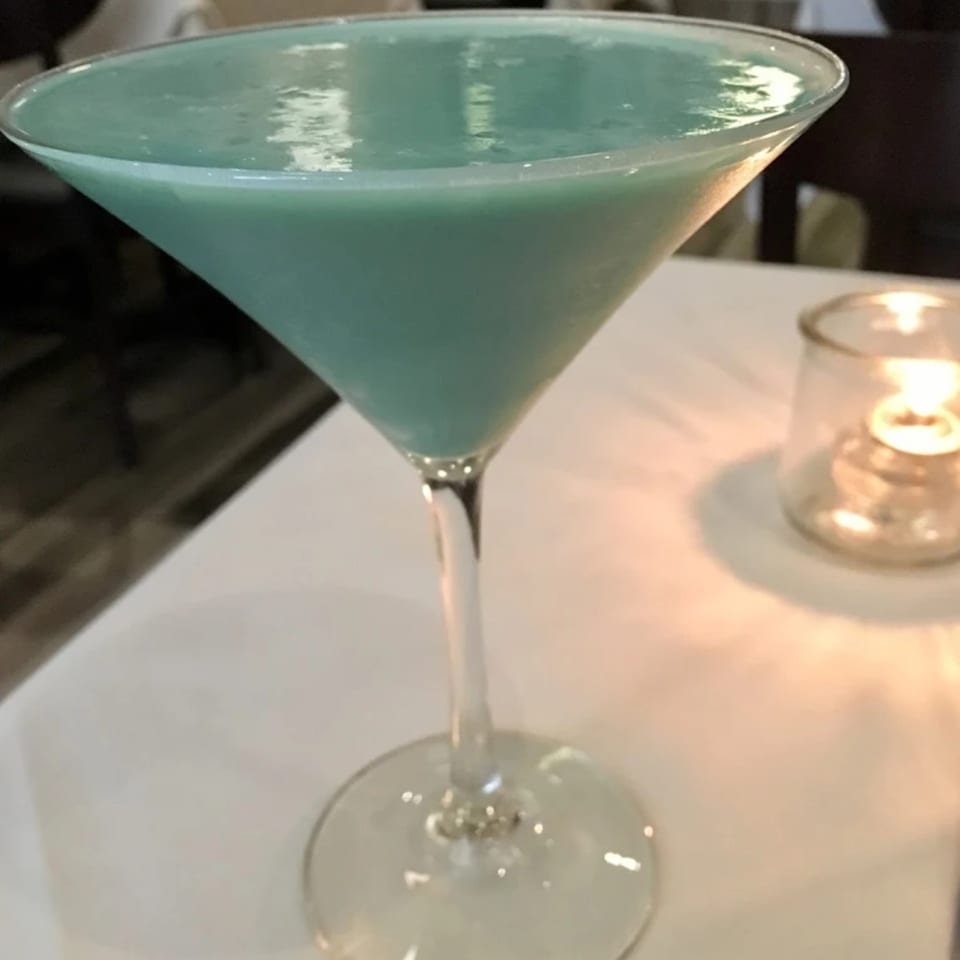 A vibrant blue-green martini in a stemmed glass sits on a white tablecloth next to a flickering candle.